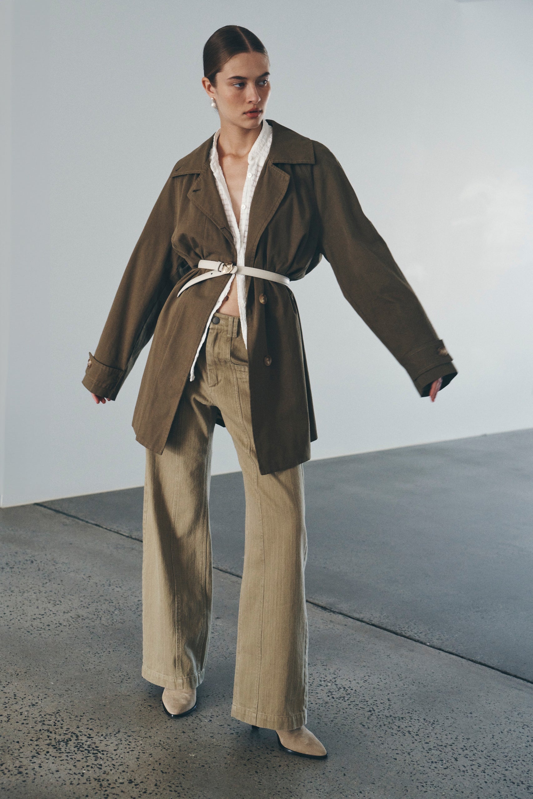 Bianca wears the Valentina Trench Jacket, Cora Blouse, and Gio Jeans. She accessorises with pearl drop earrings, a skinny white belt, and brown suede knee-high boots. Bianca completes the look with a low, slick bun. 