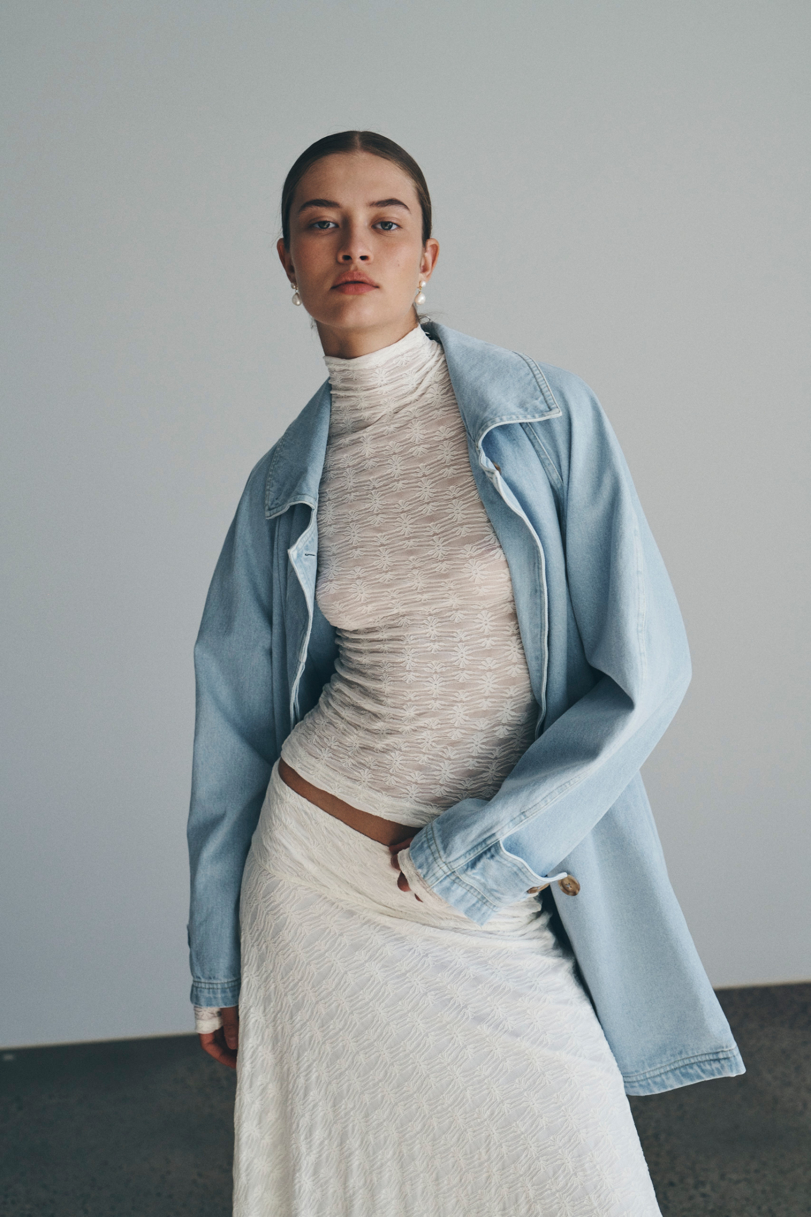 Bianca wears the Valentina Trench Jacket in Washed Denim over the Galo Flower Lace Top and Lydia Flow Lace Midi Skirt, both in Creme. She accessorises with pearl drop earrings and completes the look with a low, slick bun. 