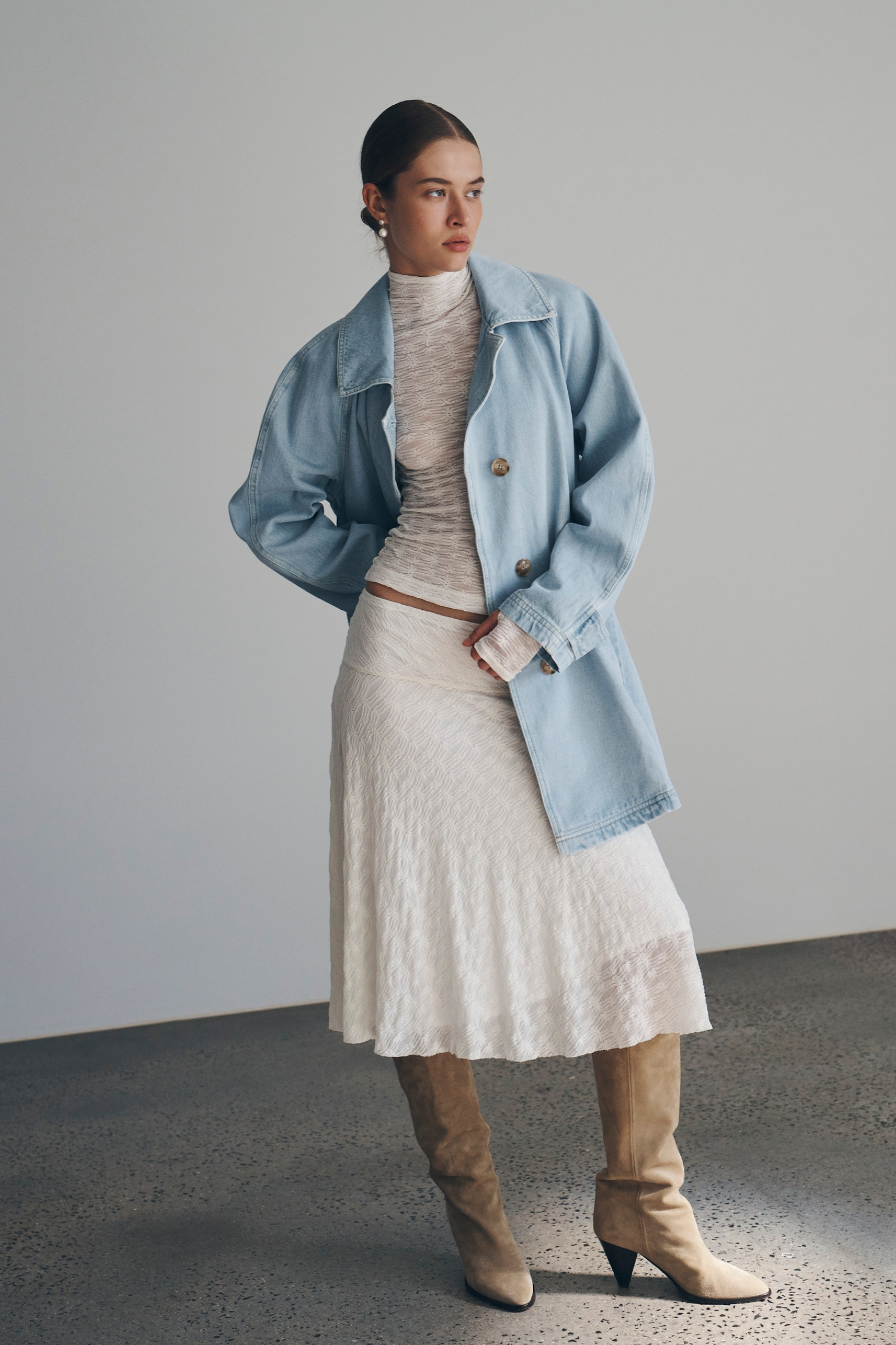 Bianca wears the Valentina Trench Jacket in Washed Denim over the Galo Flower Lace Top and Lydia Flow Lace Midi Skirt, both in Creme. She accessorises with pearl drop earrings and brown suede knee-high boots. Bianca completes the look with a low, slick bun. 