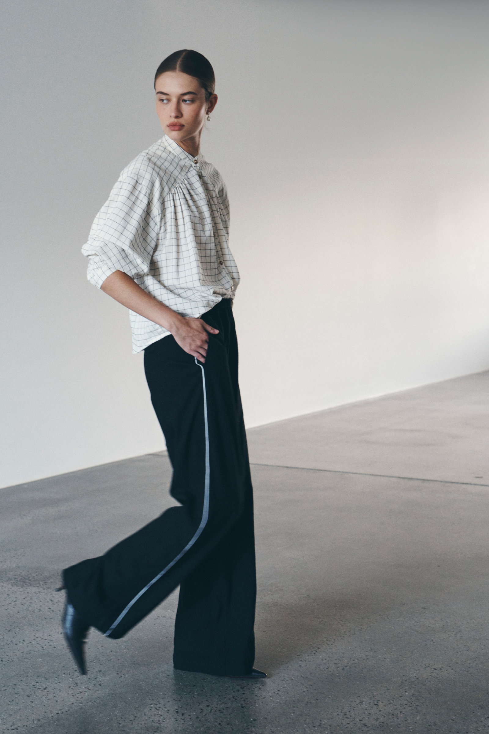 Bianca wears the Cora Blouse in Black & White Check and Vera Linen Wide Pants. She accessorises with pearl drop earrings, black leather boots, and completes the look with a low, slick bun. 