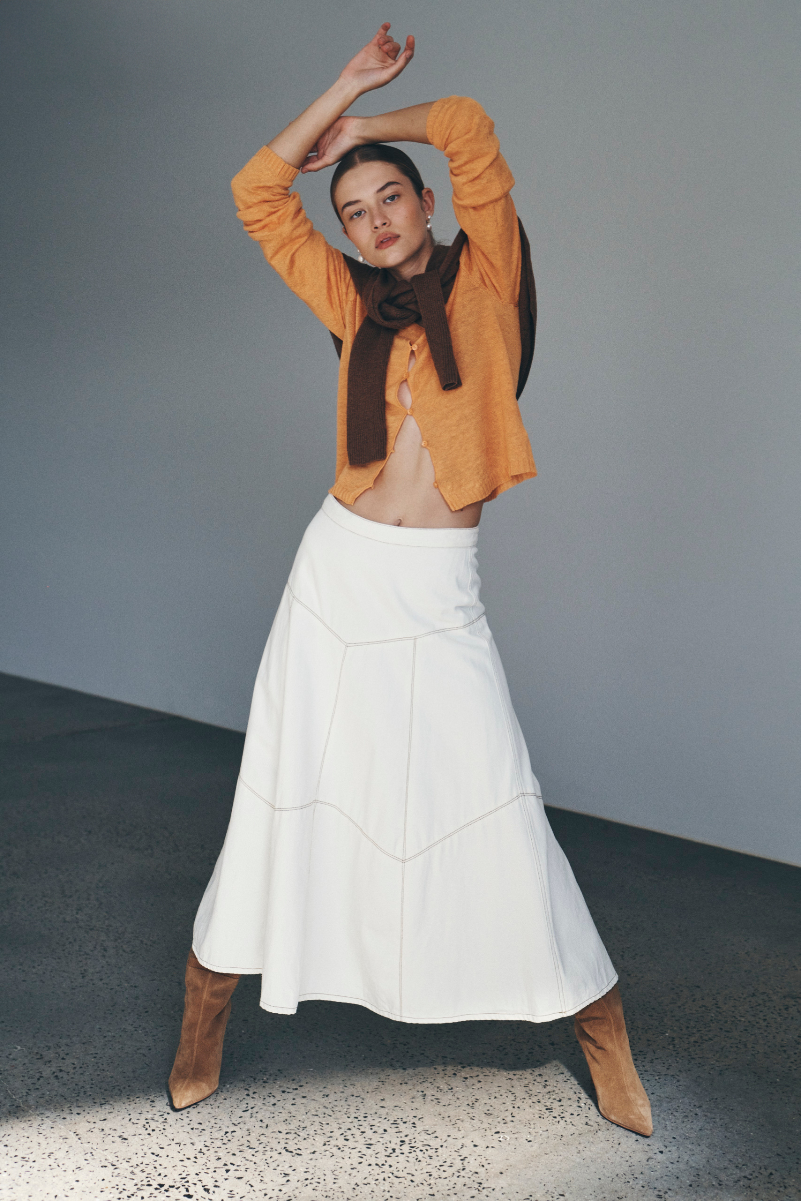 Bianca wears the Marcia Knit Long Sleeve Top and Paloma Midi Skirt with an Enzo Merino Knit Jumper tied around her shoulders. She accessorises with pearl drop earrings, brown suede knee-high boots, and completes the look with a low, slick bun. 
