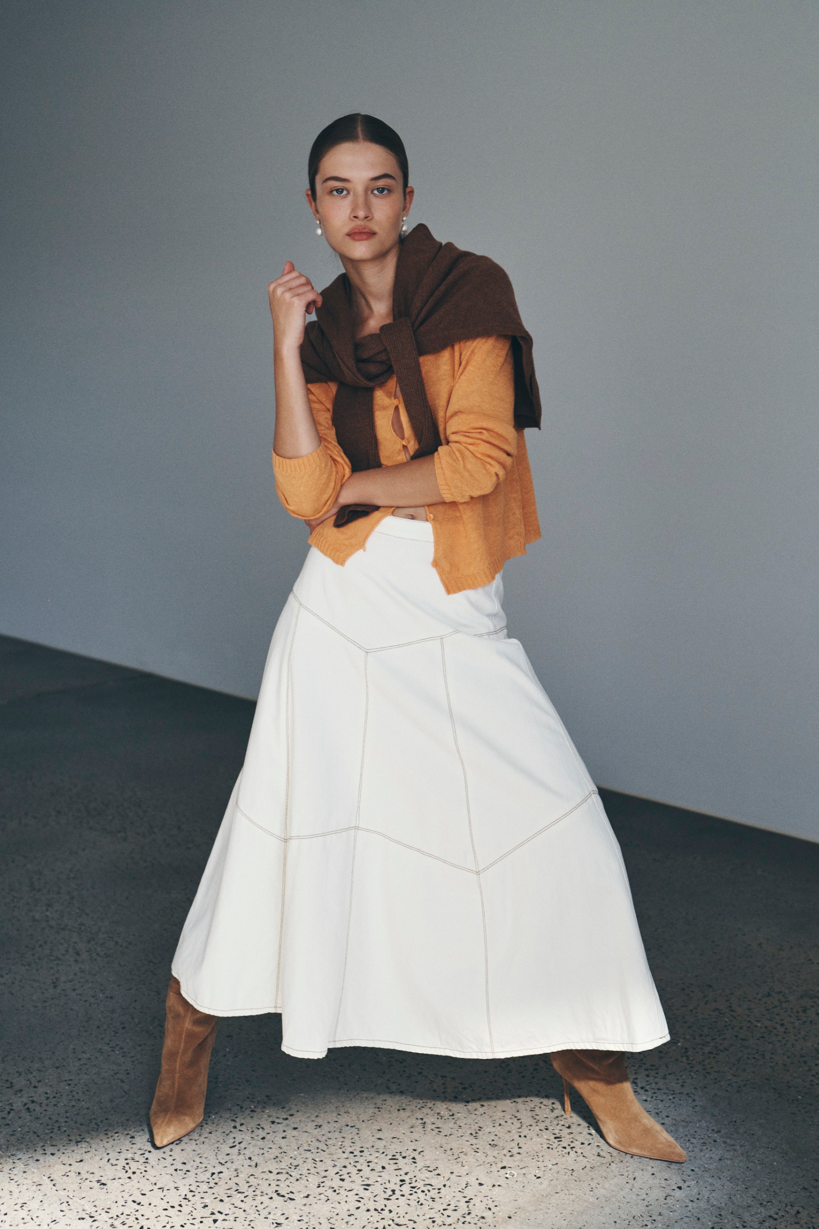 Bianca wears the Marcia Knit Long Sleeve Top and Paloma Midi Skirt with an Enzo Merino Knit Jumper tied around her shoulders. She accessorises with pearl drop earrings, brown suede knee-high boots, and completes the look with a low, slick bun. 