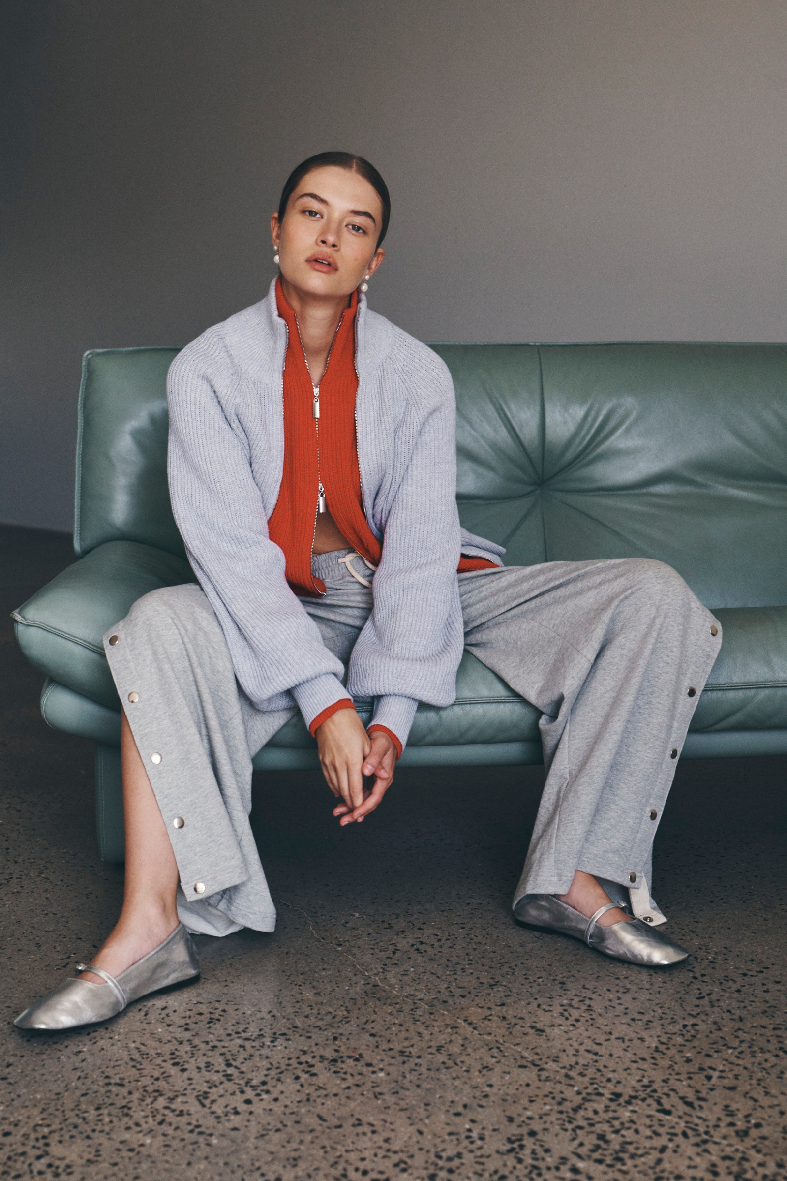 Bianca wears the Banjo Zip Knit Cardigan in both Crimson Red and Grey Marle paired with the Vena Terry Snap Pants in Grey Marle. Bianca lounges on a green leather sofa and accessorises with pearl drop earrings and silver ballet flats.