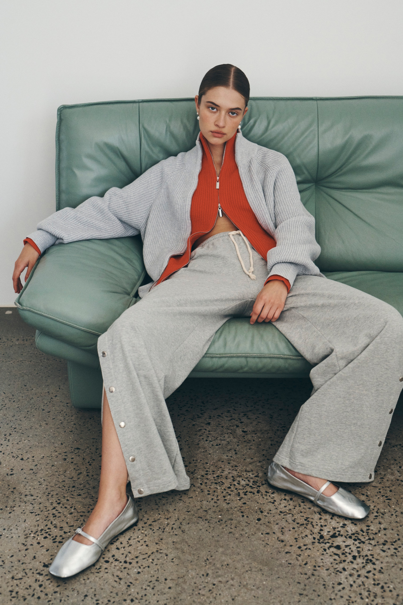 Bianca wears the Banjo Zip Knit Cardigan in both Crimson Red and Grey Marle paired with the Vena Terry Snap Pants in Grey Marle. Bianca lounges on a green leather sofa and accessorises with pearl drop earrings and silver ballet flats.