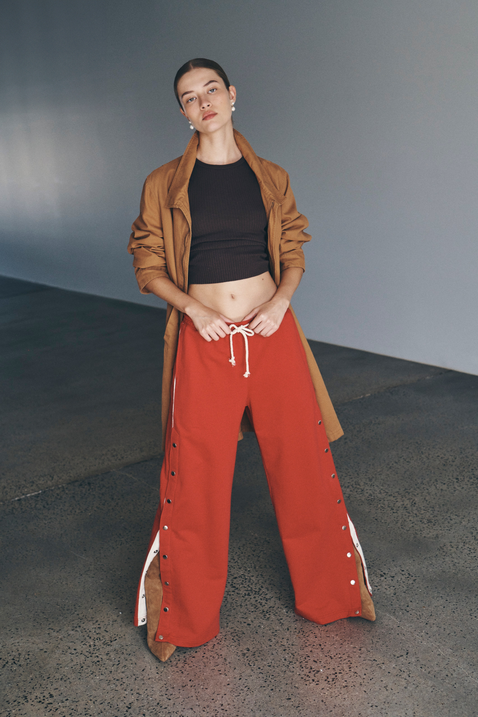 Bianca wears the Tait Jacket, Avery Knit Tank, and Vena Terry Snap Pants in Poppy Red. She accessorises with pearl drop earrings, brown suede knee-high boots, and completes the look with a low, slick bun. 