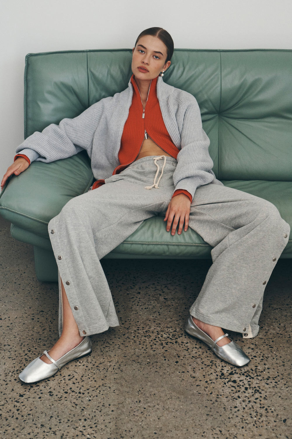 Bianca wears the Banjo Zip Knit Cardigan in both Crimson Red and Grey Marle paired with the Vena Terry Snap Pants in Grey Marle. Bianca lounges on a green leather sofa and accessorises with pearl drop earrings.
