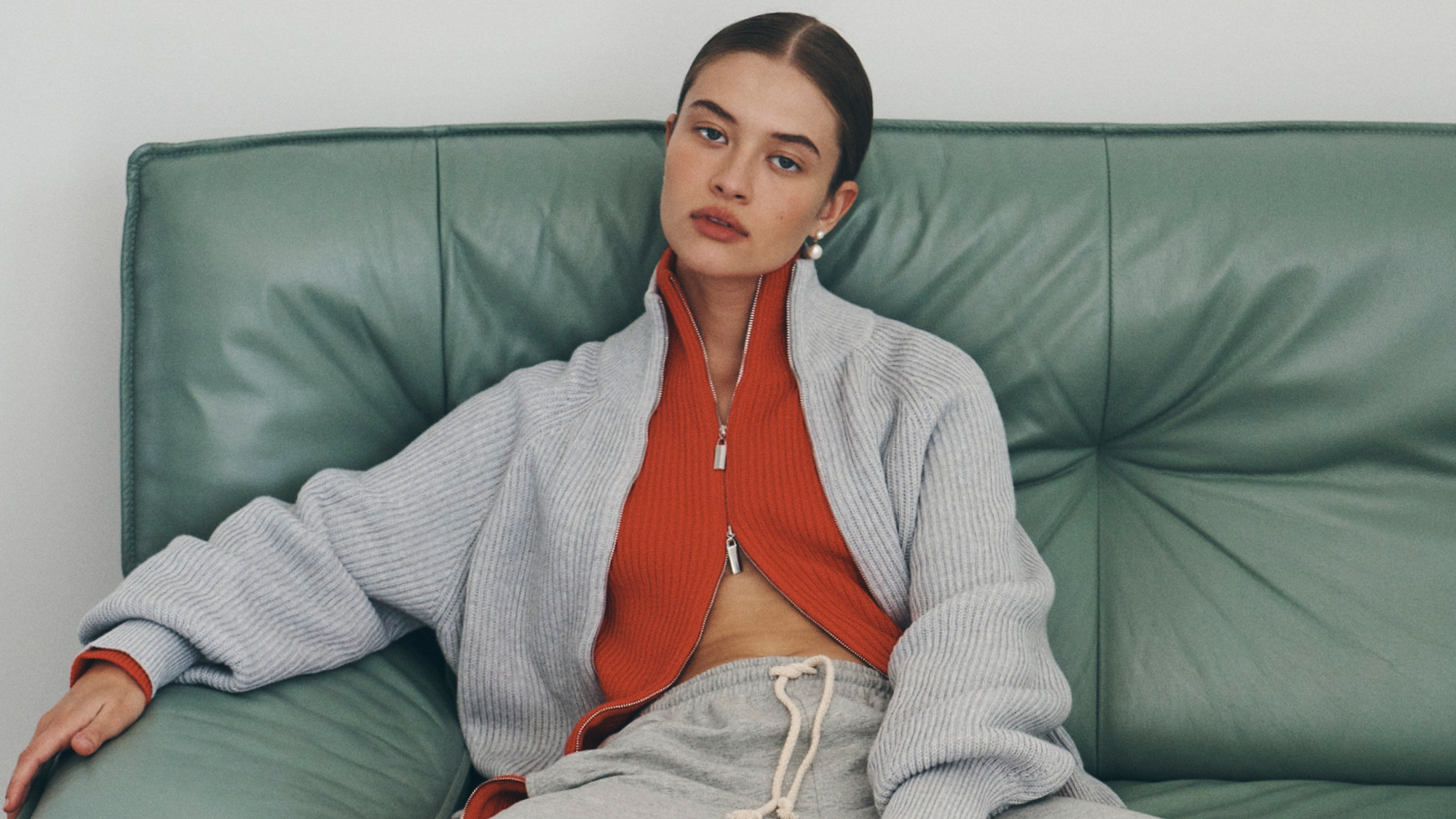 Bianca wears the Banjo Zip Knit Cardigan in both Crimson Red and Grey Marle paired with the Vena Terry Snap Pants in Grey Marle. Bianca lounges on a green leather sofa and accessorises with pearl drop earrings.